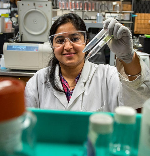 A female student smiles wearing glasses and a lab coat while holding up two test tubes